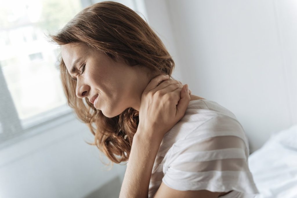 Woman sitting on her bed rubbing her neck in pain after waking up with stiffness.
